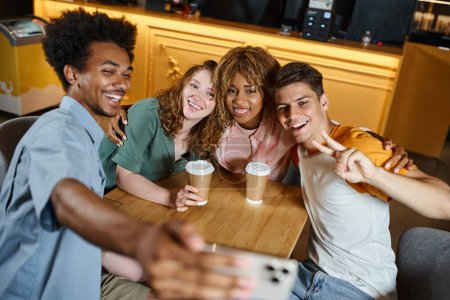 Foto de Hombre afroamericano feliz tomando selfie con los estudiantes multiétnicos de moda en la cafetería albergue, amistad - Imagen libre de derechos