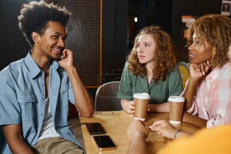 Foto de Sonriente y elegante hombre afroamericano hablando con amigos multiculturales en la cafetería del albergue de estudiantes - Imagen libre de derechos