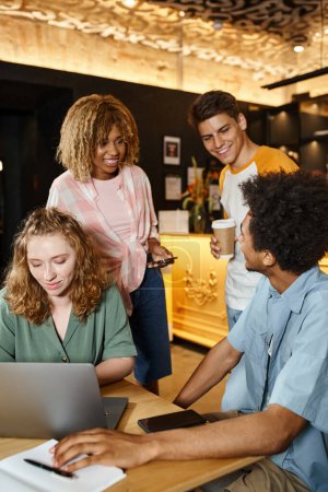 Téléchargez les photos : Jeune femme utilisant un ordinateur portable près des amis multiculturels joyeux dans le café salon de l'auberge de jeunesse - en image libre de droit