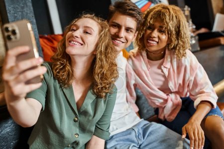 excited woman taking selfie with multiethnic buddies while having fun in lobby of students hostel