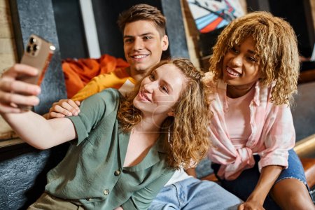 smiling woman taking selfie with carefree multiethnic students in youth hostel lounge, friendship