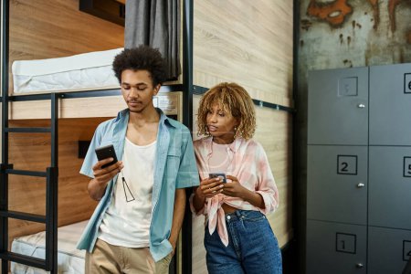 estudiantes afroamericanos de moda utilizando teléfonos inteligentes cerca de camas de dos pisos en albergue moderno