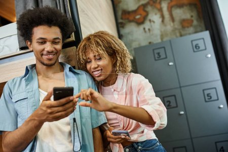 alegre pareja afroamericana mirando teléfono inteligente en acogedora habitación del albergue de estudiantes, compañeros de habitación