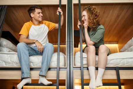 Foto de Feliz pareja de estudiantes sentados en camas de dos pisos y sonriendo el uno al otro en el albergue - Imagen libre de derechos