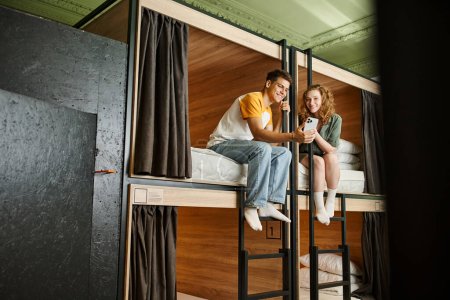 Foto de Alegre joven mujer mostrando teléfono inteligente a novio en camas de dos pisos en acogedora habitación del albergue - Imagen libre de derechos