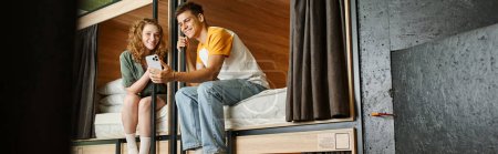 Foto de Despreocupado mujer joven mostrando teléfono inteligente a novio en camas de dos pisos en la habitación del albergue, pancarta - Imagen libre de derechos
