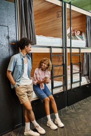 Foto de Mujer afroamericana feliz con taza de papel hablando con un amigo en la habitación del albergue con camas de dos pisos - Imagen libre de derechos