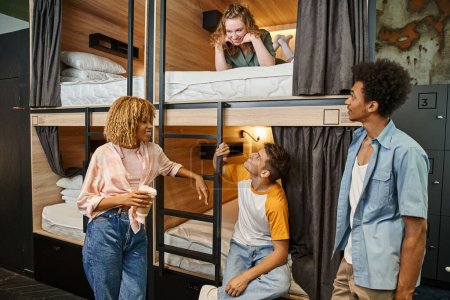 Foto de Alegres amigos multiétnicos mirando a la mujer joven acostada en la cama de dos pisos en el albergue de estudiantes - Imagen libre de derechos