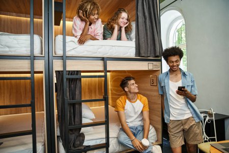 smiling african american man using smartphone near multiethnic mates on double-decker beds in hostel