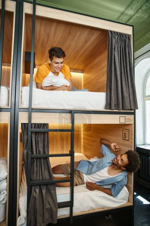 cheerful multiethnic friends lying and talking on double-decker beds in cozy room of students hostel