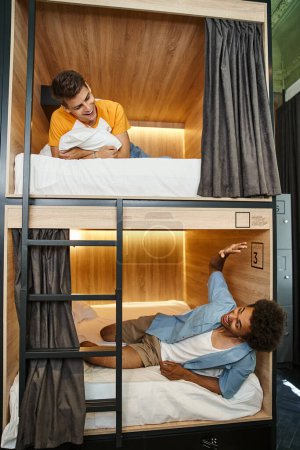 excited african american student lying on double-decker bed and waving hand to friend in hostel room