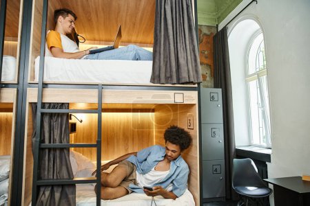 interracial friends with laptop and smartphone on double-decker beds in modern hostel, vacation