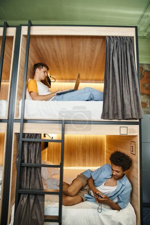 Foto de Estudiantes multiétnicos que utilizan el ordenador portátil y teléfono inteligente en camas de dos pisos en el acogedor albergue juvenil - Imagen libre de derechos