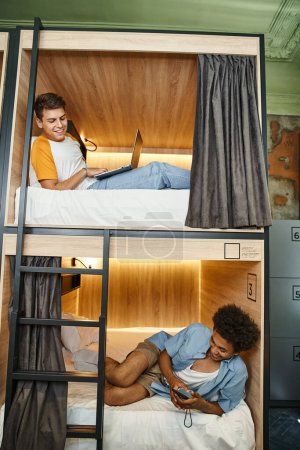 joyful multiethnic roommates lying with laptop and smartphone on double-deckers beds in hostel