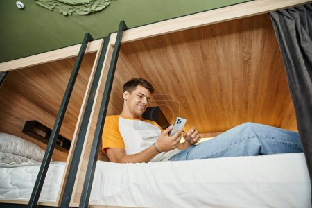 Foto de Vista de ángulo bajo de hombre joven navegar por Internet en el teléfono inteligente en la cama de dos pisos en el albergue - Imagen libre de derechos