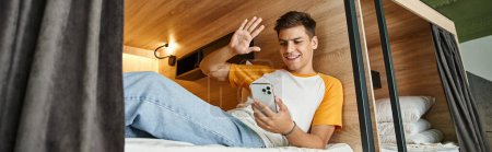 alegre chico saludando la mano durante la videollamada en cama de dos pisos en acogedor albergue de estudiantes, pancarta