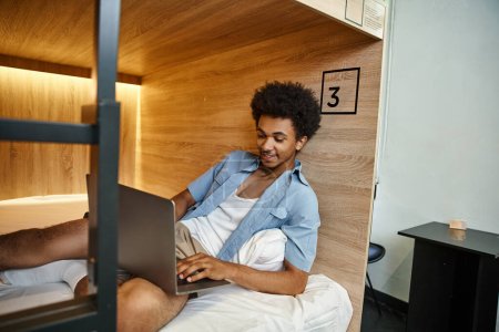 Foto de Feliz y elegante afroamericano estudiante de networking en el ordenador portátil en la cama de dos pisos en el albergue - Imagen libre de derechos