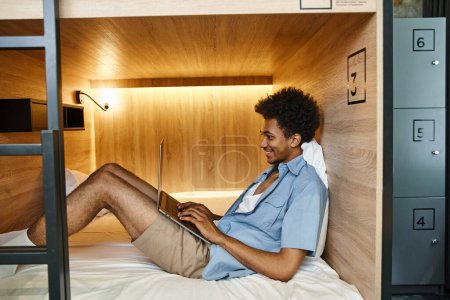 Foto de Vista lateral del hombre americano africano alegre mecanografiando en el ordenador portátil en la cama acogedora de dos pisos en el albergue - Imagen libre de derechos
