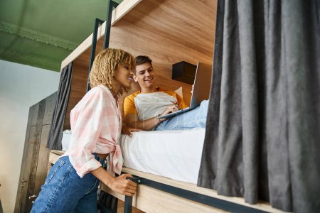 cheerful african american woman near friend using laptop on double-decker bed in students hostel