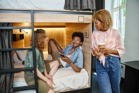 cheerful african man with smartphone on double-decker bed near interracial female friends in hostel