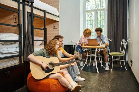 Téléchargez les photos : Étudiants multiculturels avec guitare et ordinateur portable dans la chambre de l'auberge de jeunesse contemporaine, les voyageurs - en image libre de droit