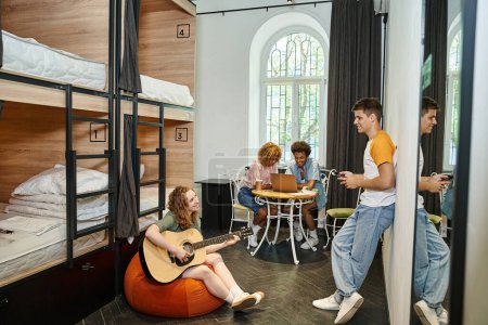 multiethnic buddies with guitar and laptop in room of cozy youth hostel, friendship and travel