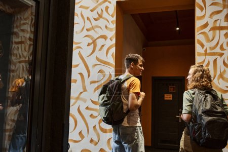 Téléchargez les photos : Vue arrière de sourire couple d'étudiants parlant dans le couloir de l'auberge de jeunesse avec un design moderne - en image libre de droit