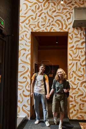 happy young couple holding hands and looking at camera in corridor of hostel with modern design