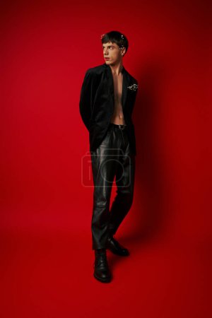 Photo for Young man in stylish outfit with flowers in hair and pocket of velvet blazer posing on red backdrop - Royalty Free Image