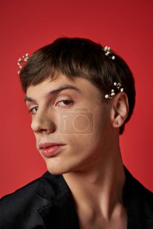 good looking young man in stylish outfit with flowers in hair looking at camera on red background