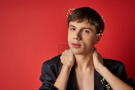 jeune homme en velours blazer avec des fleurs dans les cheveux en regardant la caméra et toucher le cou sur fond rouge