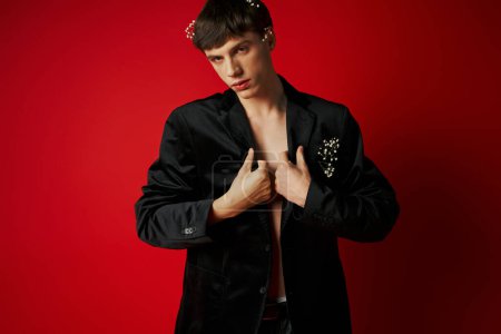 elegant young man in velvet blazer with flowers in hair looking at camera and posing on red backdrop