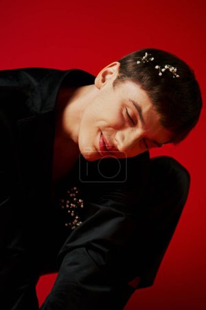 Téléchargez les photos : Joyeux jeune homme en velours noir veste avec des fleurs dans les cheveux souriant sur fond rouge, glamour - en image libre de droit