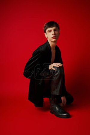 Photo for Young man in stylish black attire with leather pants and flowers in hair sitting on red background - Royalty Free Image