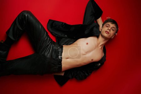 top view of young man in jacket and leather pants lying and looking at camera on red background
