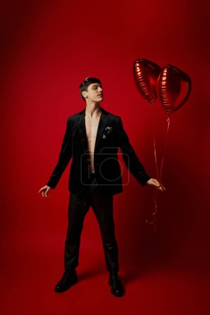 Foto de Longitud completa de joven guapo en traje negro sosteniendo globos en forma de corazón sobre fondo rojo - Imagen libre de derechos