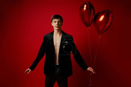 glücklicher und gutaussehender junger Mann in schwarzer Kleidung mit herzförmigen Luftballons auf rotem Hintergrund