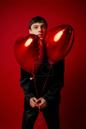 schöner junger Herr in schwarzer Kleidung mit herzförmigen Luftballons auf rotem Hintergrund
