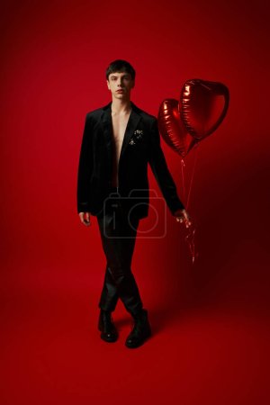 full length of stylish male model in black attire holding heart-shaped balloons on red backdrop