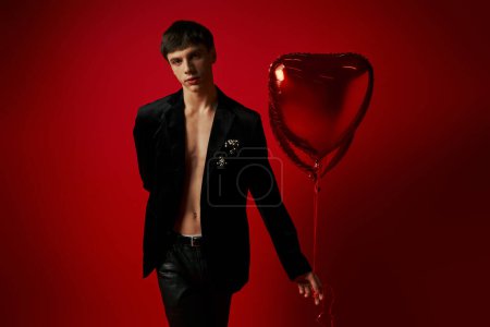 stylish male model in velvet jacket and leather pants holding heart-shaped balloons on red backdrop