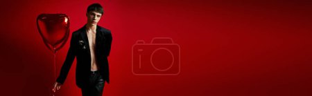 stylish man in velvet jacket and leather pants holding heart-shaped balloons on red backdrop, banner
