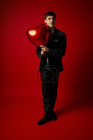 happy male model in velvet jacket and leather pants holding heart-shaped balloon on red backdrop