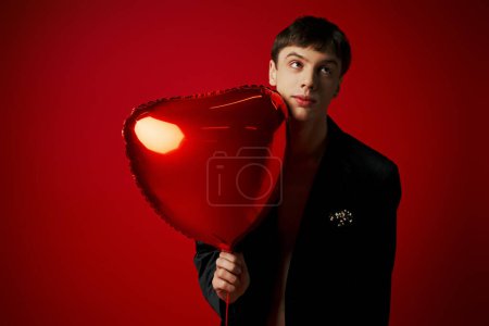 confused young man in velvet jacket holding heart-shaped balloon and looking away on red background