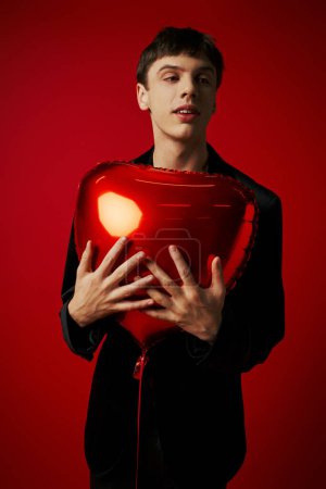 young male model in velvet jacket and leather pants holding heart-shaped balloon on red backdrop
