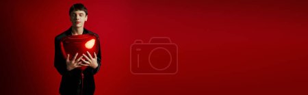 Téléchargez les photos : Heureux gen z gars en velours veste souriant et tenant ballon en forme de coeur sur fond rouge, bannière - en image libre de droit