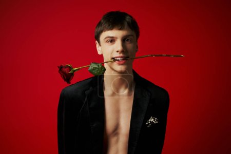 romantic young man in velvet jacket holding rose in teeth and smiling on red background, flirt