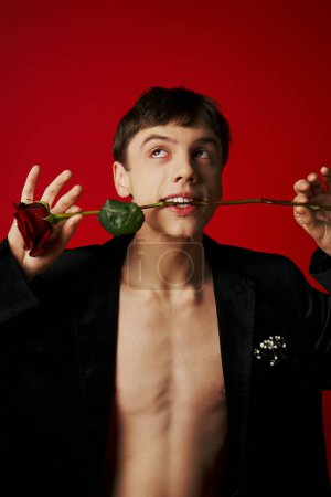 romantic young man in velvet jacket holding rose in teeth and looking up on red background, flirt