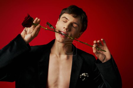 shirtless young man with closed eyes posing in velvet jacket holding rose in teeth on red background