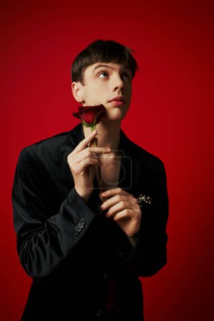 romantic guy in velvet jacket holding rose in hands and looking up on red background, flirt