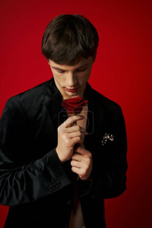 romantischer und eleganter junger Mann in Samtjacke mit Rose in der Hand auf rotem Hintergrund, Flirt